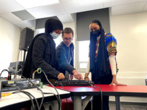 Three adults (a woman wearing a veil and a mask, a white man with glasses, and a young Black woman with a mask)stand around a sound mixing table.